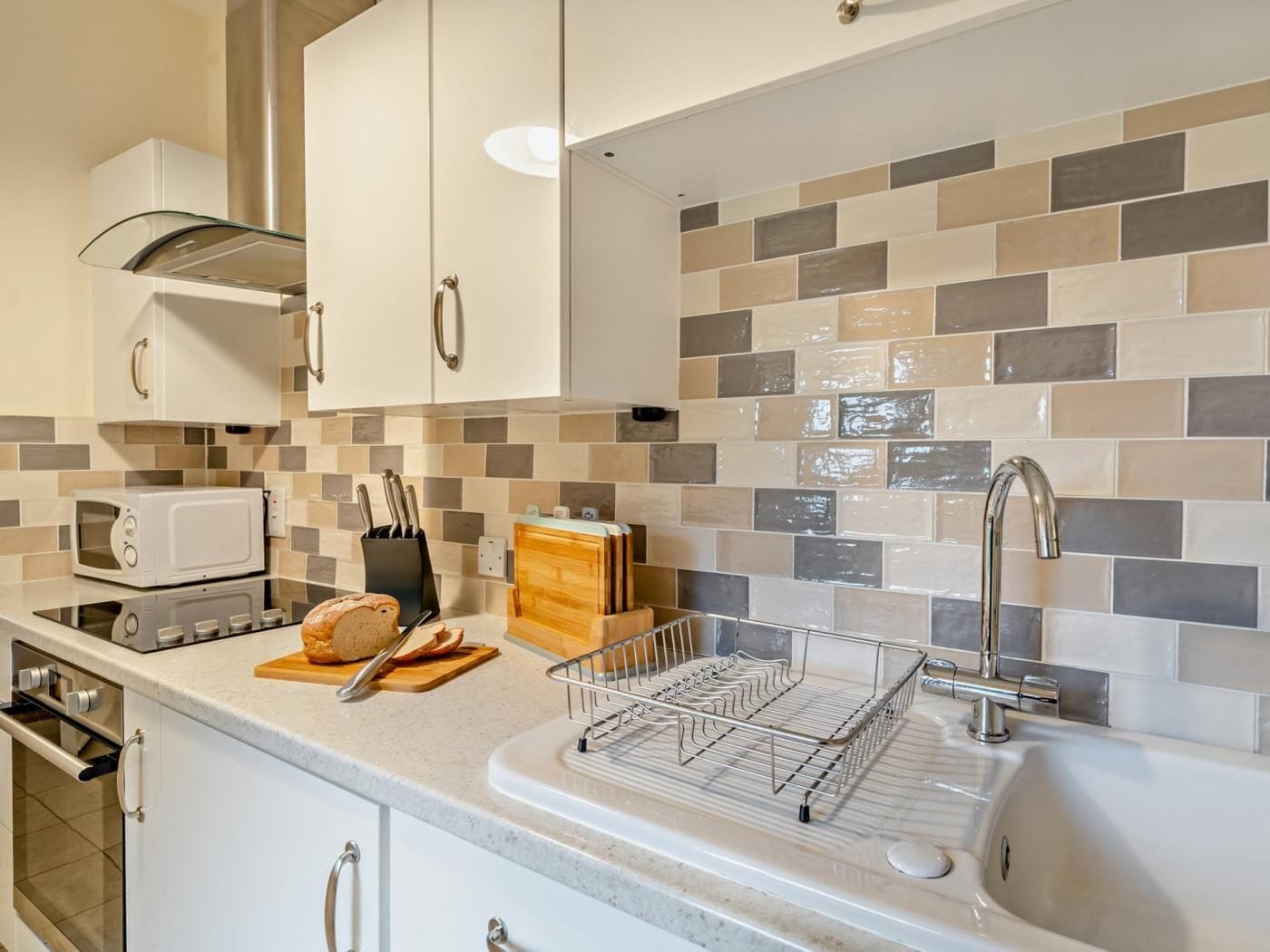 Cozinha com armários brancos, micro-ondas, torradeira, pia com torneira moderna e painel de azulejos.