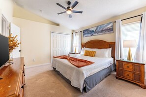 Une chambre à coucher comprenant un lit, une table de chevet, une commode, un ventilateur de plafond et un tableau accroché au mur.