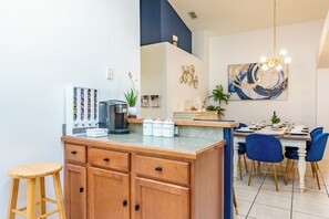 Une cuisine moderne équipée d’une machine à café, de récipients pour le thé et le café, et d’un coin repas avec des chaises bleues et une table dressée.
