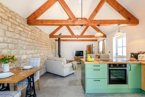 A rustic kitchen with a stone wall, wooden beams, a green countertop, and a dining area with a table set for two.