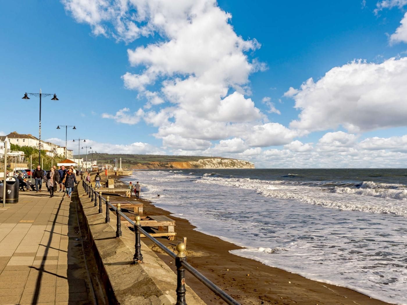 A coastal promenade with a stone walkway, wooden railings, and a view of the sea and cliffs.