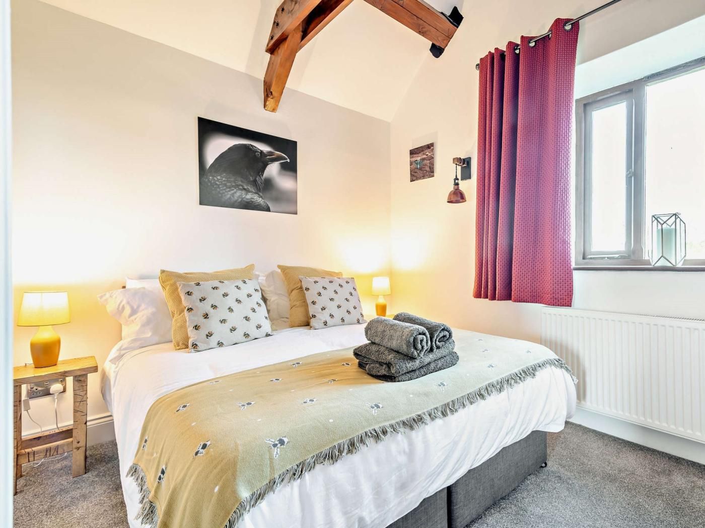 Une chambre à coucher avec un lit, des oreillers, une couverture, une fenêtre avec des rideaux, un tableau accroché au mur et une table de chevet avec une lampe.