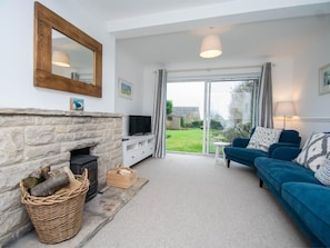 A living room with a stone fireplace, a television, a blue sofa, and a view of the outdoors through a glass door. - 3 Bed in Swanage (oc-dc054) (Worth Matravers)