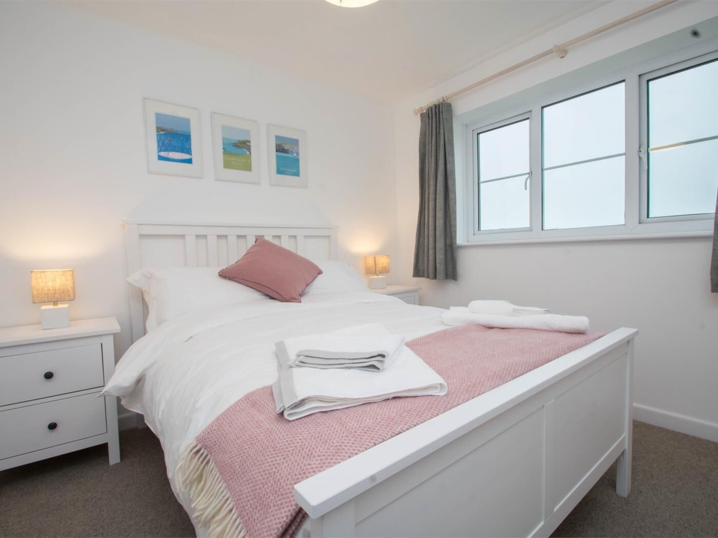 A neatly made bed with pillows and folded towels, a bedside lamp, a nightstand with a drawer, and a window with curtains.