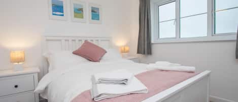 A neatly made bed with pillows and folded towels, a bedside lamp, a nightstand with a drawer, and a window with curtains.