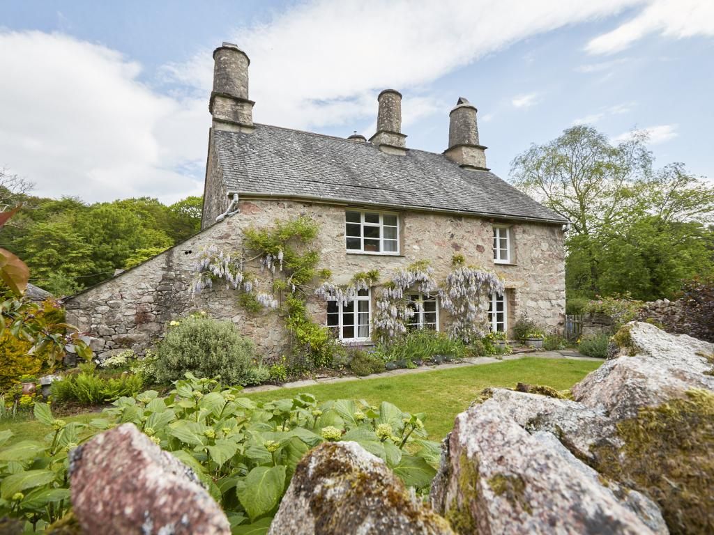 A stone house with a sloped roof and chimneys, surrounded by a well-maintained garden with large rocks in the foreground.