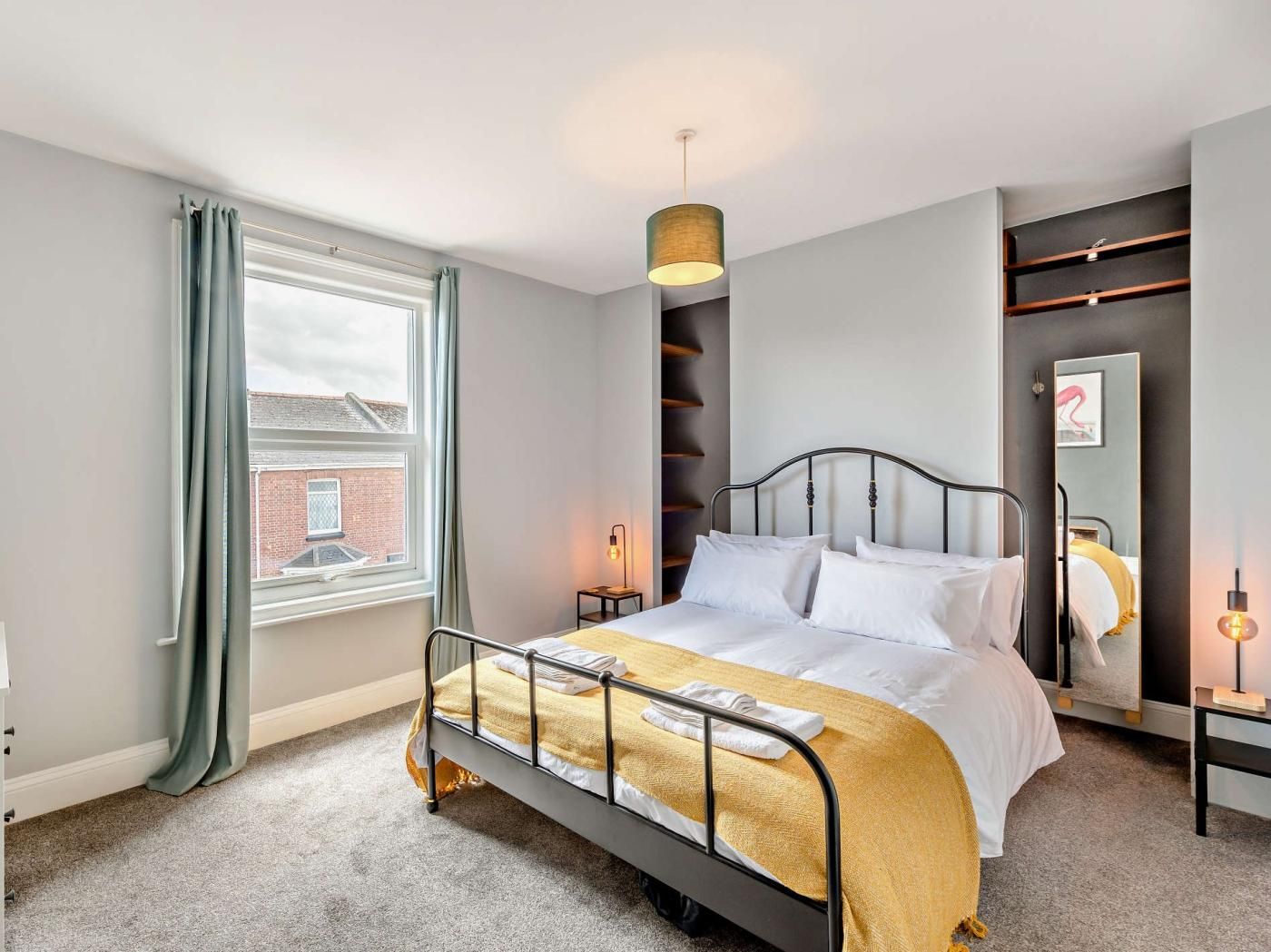 A modern hotel room with a bed, a window with curtains, a wall-mounted light, and a mirror.