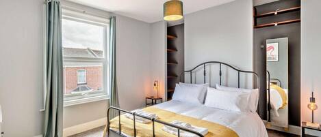 A modern hotel room with a bed, a window with curtains, a wall-mounted light, and a mirror.
