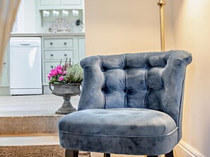 A room with a tufted armchair, a white kitchen area with a dishwasher, and a potted plant with pink flowers. - 2 Bed in Cockermouth (oc-93280) (Bridekirk)