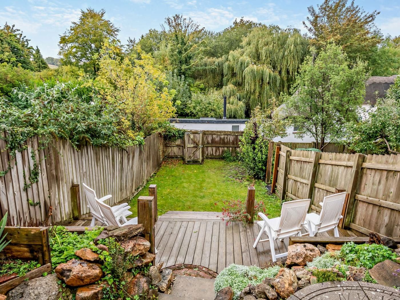 Een achtertuin met een houten terras, witte stoelen en een tuin met rotsen en planten.