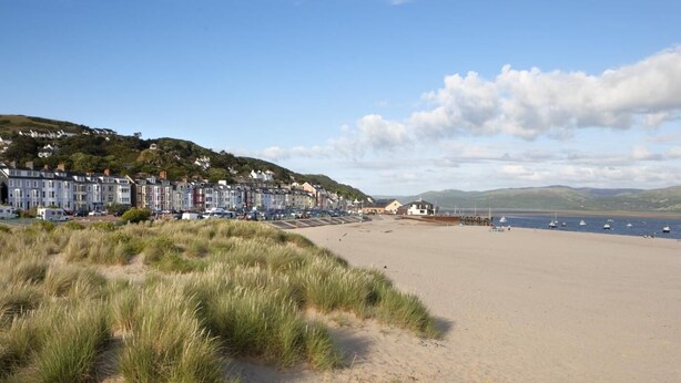 Ein Küstenstrand mit einer Reihe terrassenförmig angelegter Häuser, Sandstrand und Booten im Wasser.