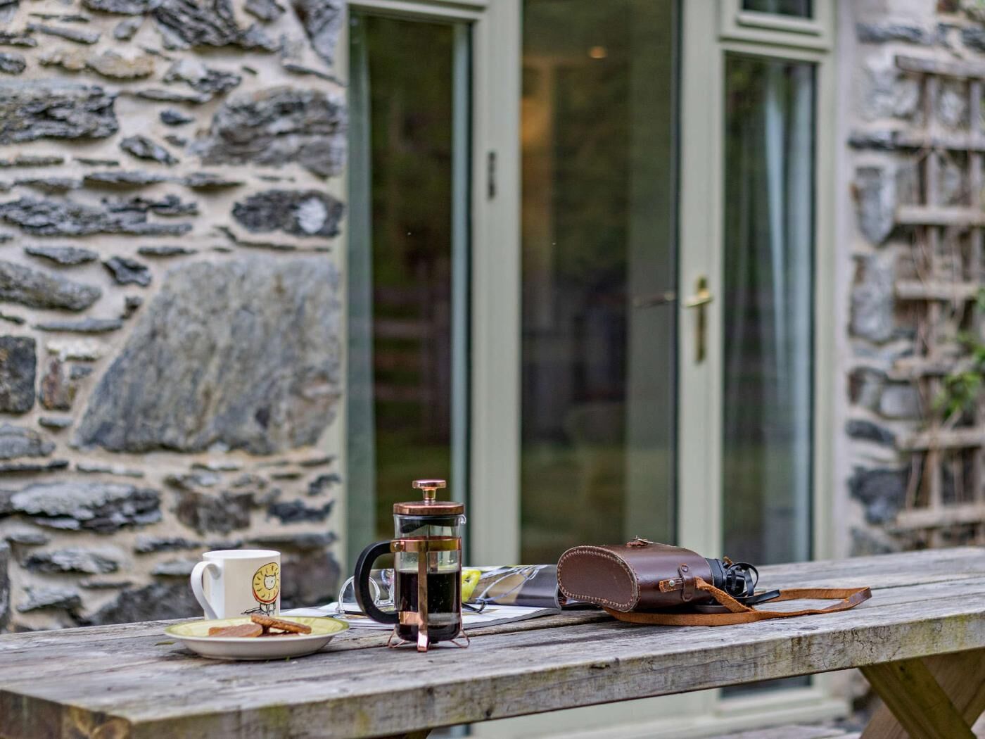 Un mur de pierre, une table en bois avec une cafetière, une tasse et une assiette remplie de nourriture.