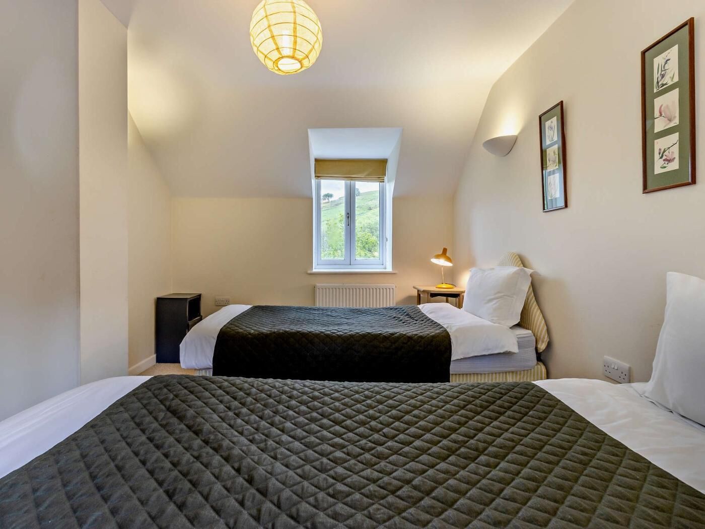 Une chambre avec deux lits, une fenêtre, une lampe et des œuvres d’art encadrées accrochées au mur.