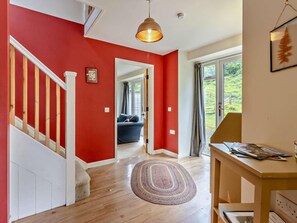 A hallway with a wooden staircase, a red wall, a blue sofa in the background, and a table with magazines. - 2 Bed in Machynlleth (oc-93645) (Glaspwll)