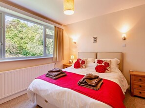 A hotel room with a large bed, two bedside lamps, a window with curtains, a radiator, and a small wooden nightstand. - 4 Bed in Peterborough (oc-93450) (Maxey)