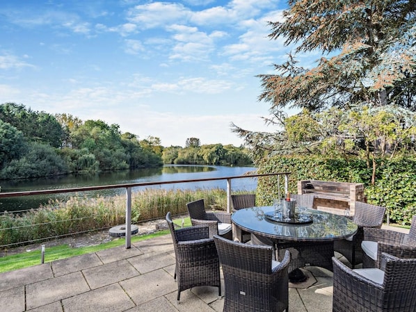 An outdoor patio with wicker furniture and a glass table, overlooking a river and surrounded by greenery.