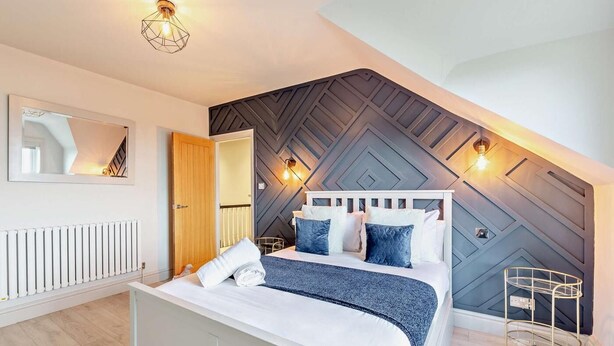 A modern bedroom with a bed, a radiator, a mirror, and a wall with geometric patterns. - 5 Bed in Aberdovey (oc-93603) (Aberdovey)