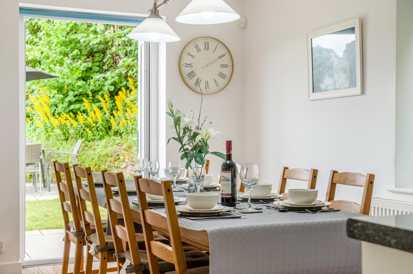 A dining area with a table set for four, a clock on the wall, and a window with a view of greenery.