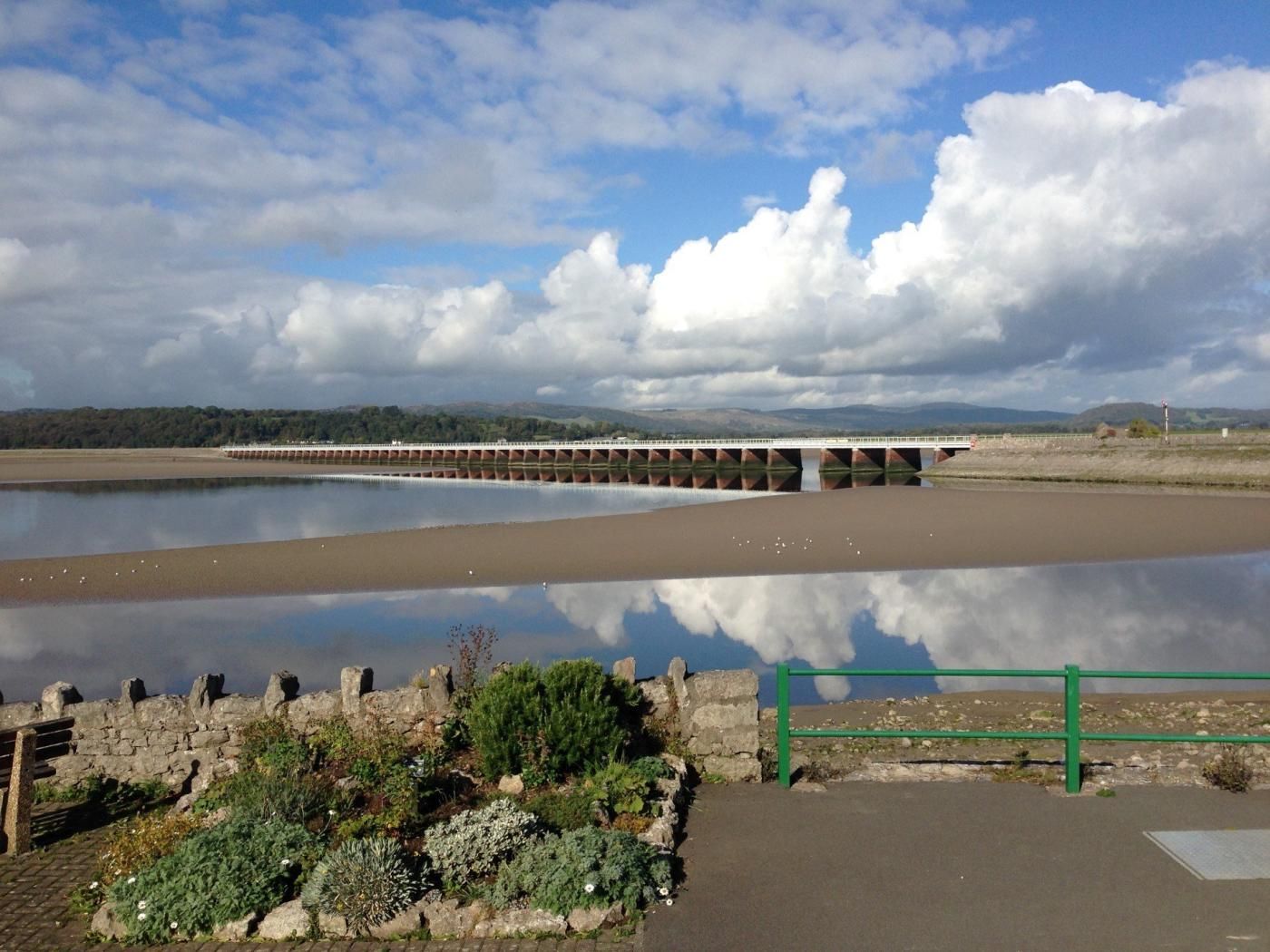 A bridge over a calm river with a stone wall and greenery in the foreground.