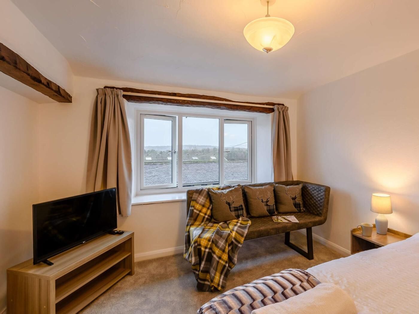 A bedroom with a bed, a TV on a wooden stand, a bench with a plaid blanket, a window with curtains, and a lamp on a bedside table.