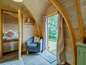 Une pièce chaleureuse aux murs en bois, avec un lit orné d’un coussin à rayures, un fauteuil bleu et une vue sur la verdure à l’extérieur.