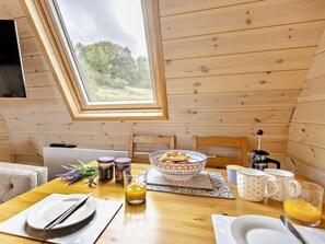 A breakfast setup in a wooden cabin with a table set for two, including plates, cutlery, a bowl of food, a coffee pot, and a glass of orange juice.