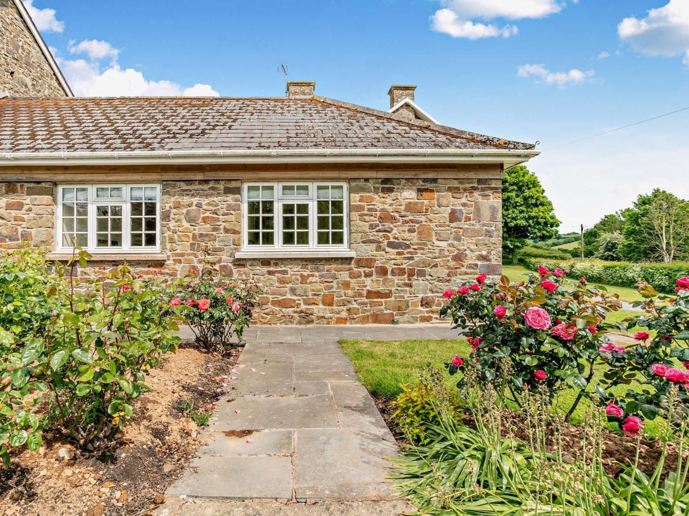 A stone house with two windows, a garden with roses, and a clear sky.