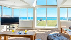 Ein Wohnzimmer mit Fernseher, Couchtisch mit Getränken, Ledersofas und großen Fenstern mit Meerblick.