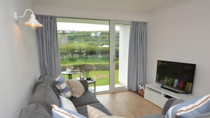 A modern living room with a sofa, TV, and a view of a green landscape through a sliding door. - 2 Bed in Newquay (oc-43204) (Mawgan Porth)