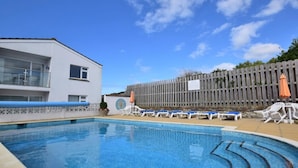 A swimming pool with lounge chairs and umbrellas, a white building with balconies, and a wooden fence. - 2 Bed in Newquay (oc-43204) (Mawgan Porth)