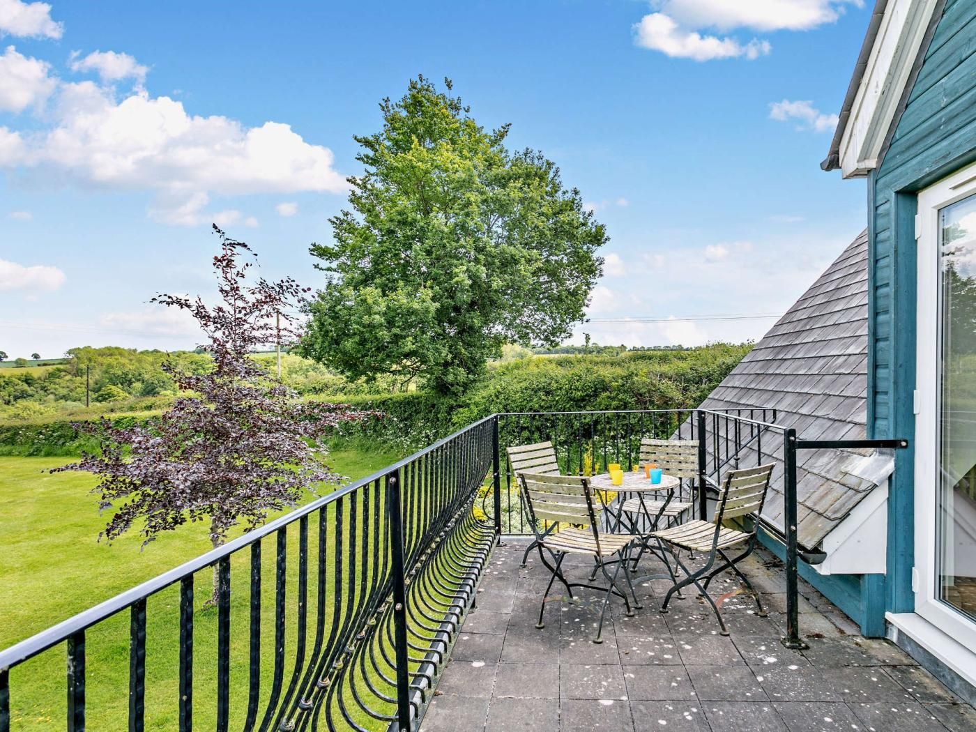A balcony with a view of a green landscape and a tree, featuring outdoor furniture and a railing.