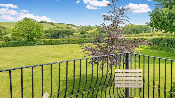A balcony with a wooden table, metal railing, and a view of a green landscape. - 3 Bed in Crediton (oc-waie2) (Zeal Monachorum)