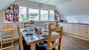 A dining area with a wooden table set for four, a window with curtains, a clock, and a view of greenery outside. - 3 Bed in Crediton (oc-waie2) (Zeal Monachorum)