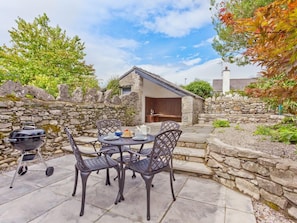 A stone-walled outdoor patio with a round table and chairs, a barbecue grill, and a stone building in the background. - 1 Bed in Grange-over-sands (oc-78659) (Cartmel)