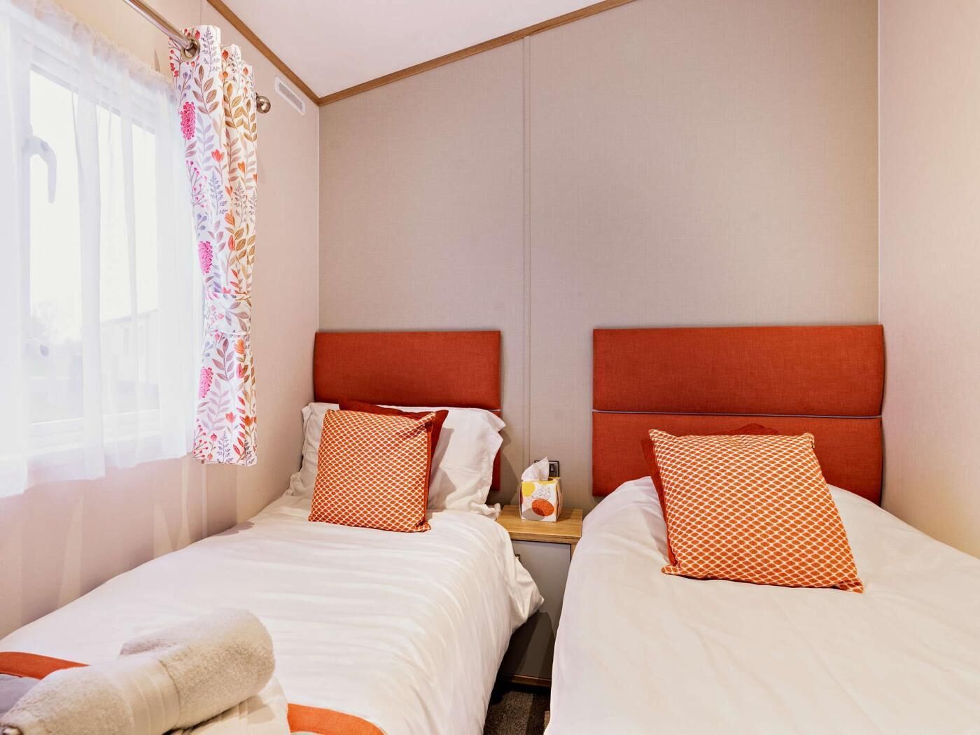 A hotel room with two beds, each with a red headboard, white bedding, and patterned pillows. A window with sheer curtains and a floral pattern is visible.