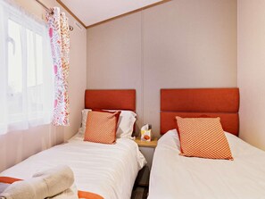 Quarto de hotel com duas camas, cada uma com cabeceira vermelha, roupa de cama branca e almofadas estampadas. Uma janela com cortinas leves e padrão floral é visível.