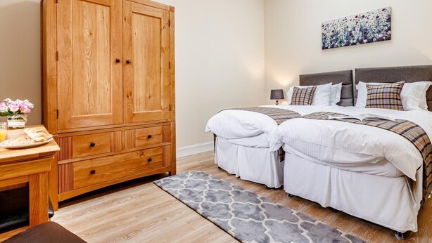 Une chambre avec une grande armoire en bois, un lit avec une literie blanche, un tapis à motifs gris et un tableau accroché au mur.