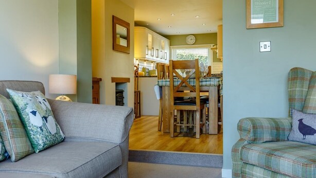 Ein Wohnzimmer mit einer Couch, einem karierten Sessel, einem Esstisch mit Stühlen und einer Küche im Hintergrund.
