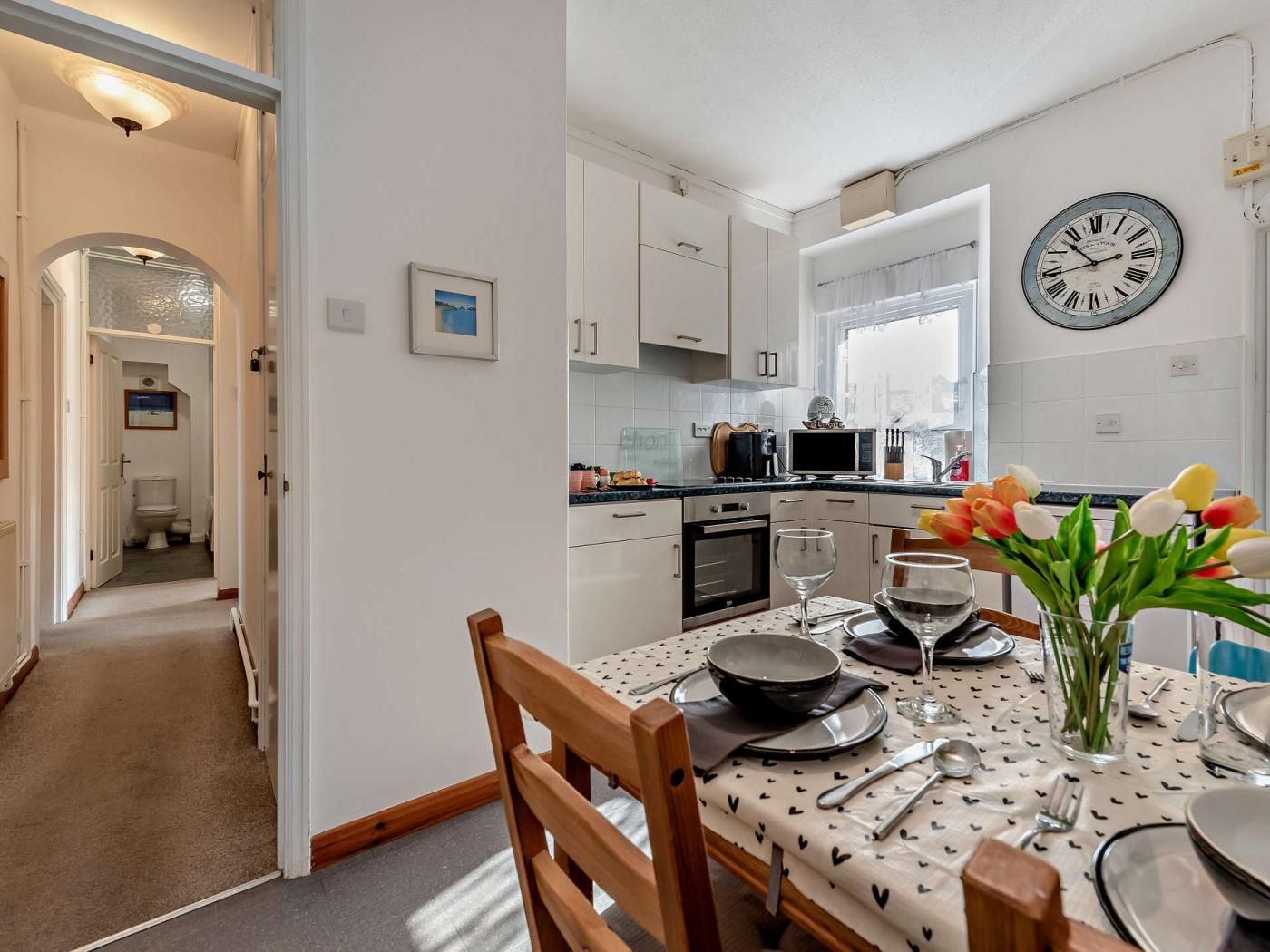 A modern kitchen with white cabinets, a clock, and a dining area set for two with a table set for a meal.