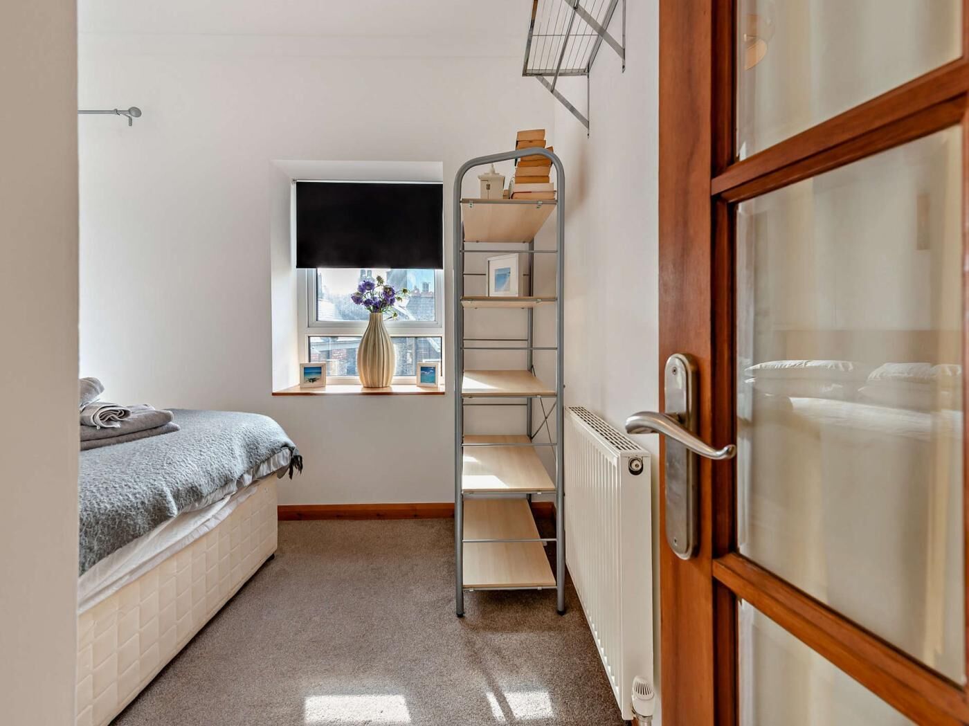 Une petite pièce, de taille modeste, comprenant un lit, un radiateur, une fenêtre donnant sur un paysage agréable et une étagère en métal.