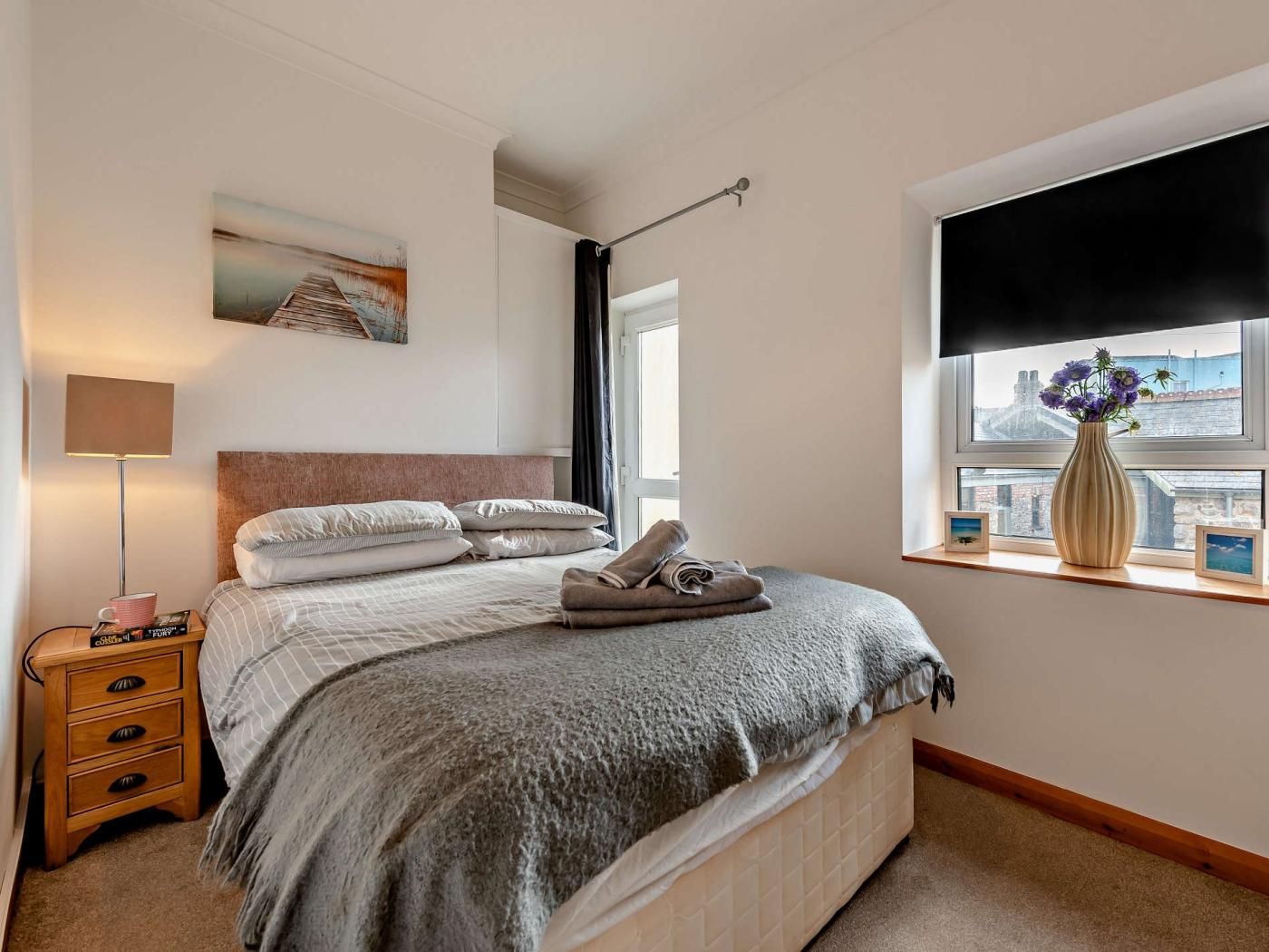 Un dormitorio ordenado con cama, mesita de noche, lámpara y ventana con vistas.