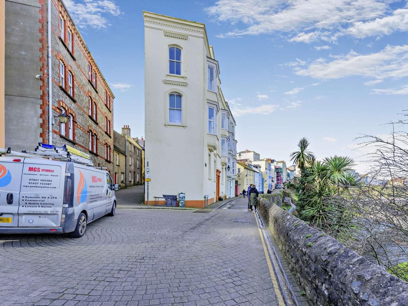 A narrow street lined with colorful buildings, a parked van, and a stone wall on one side.