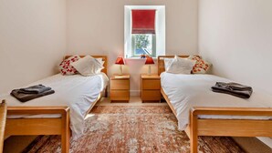 A bedroom with two beds, a window with a red blind, wooden headboard, a small bedside table with a lamp, and a patterned rug. - 2 Bed in Honiton (oc-93402) (Luppitt)
