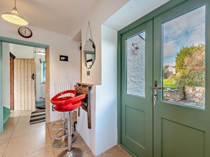 A hallway with a green door, a red stool, a clock, and a view of greenery outside.