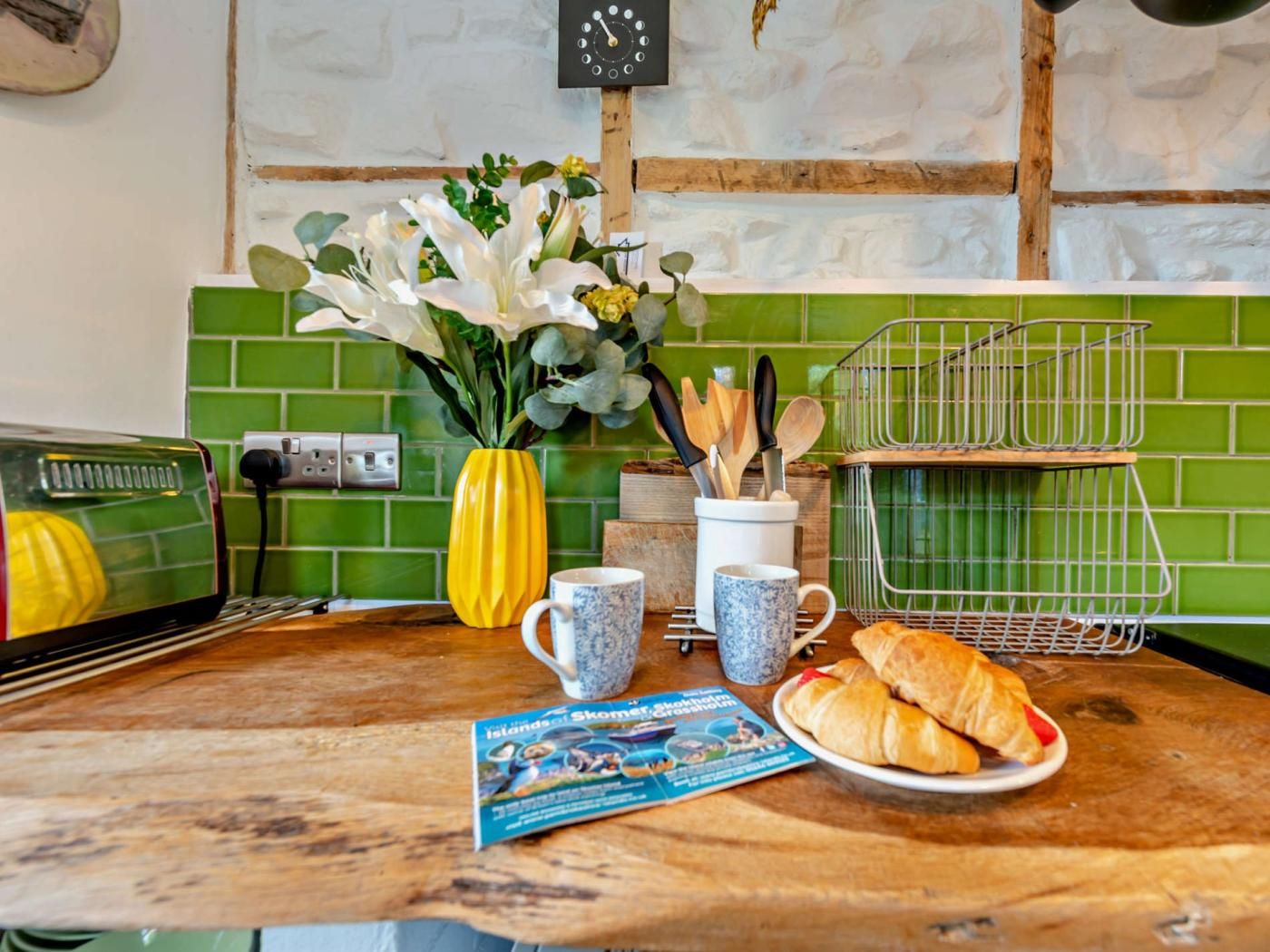 Un comptoir de cuisine avec un grille-pain, un vase rempli de fleurs, un magazine, une assiette de croissants et une horloge au mur.