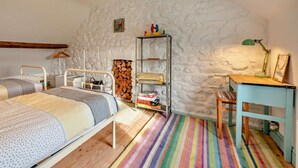 A bedroom with two beds, a wooden floor, a colorful striped rug, a stone wall, a metal shelf with books, and a desk with a lamp. - 2 Bed in Haverfordwest (oc-93577) (Marloes)