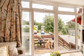 A room with a large sliding glass door leading to an outdoor deck with a dog, a bench, and a table with a vase of flowers. - 2 Bed in Nolton Haven (oc-fb098) (Nolton Haven)