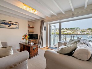 Ein Wohnzimmer mit Fernseher, einem Holztisch und einer Couch. Ein Fenster bietet Blick auf einen Hafen und Gebäude.