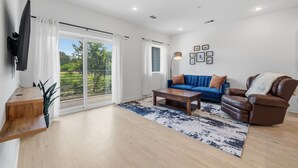 A spacious living room with a large sliding glass door, a blue sofa, a brown leather chair, a wooden coffee table, and a black floor lamp. - 2BR South Congress Condo for 6, Near DT! (Austin)