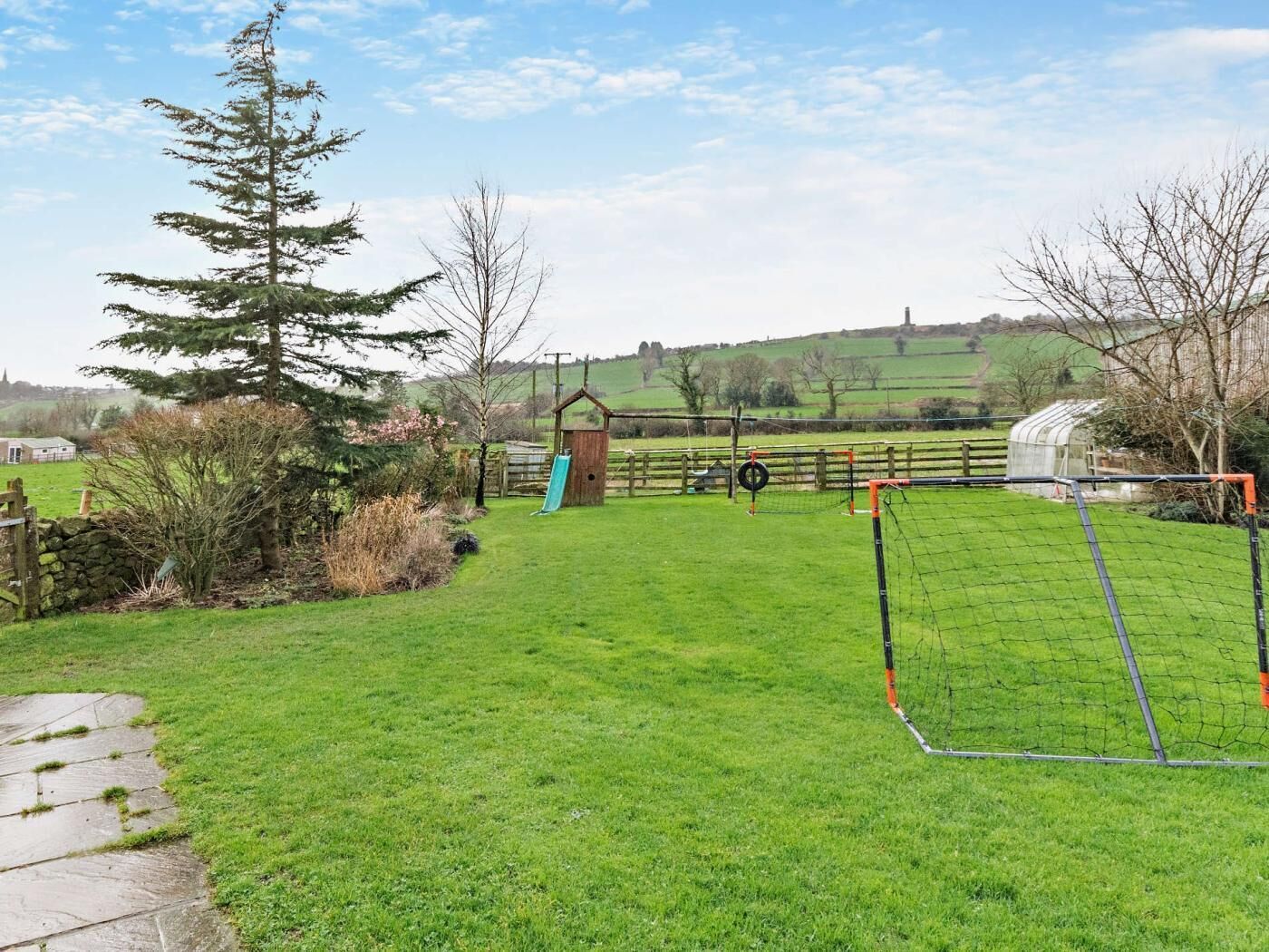 A grassy outdoor area with a small wooden structure, a fenced play area, and a stone wall.
